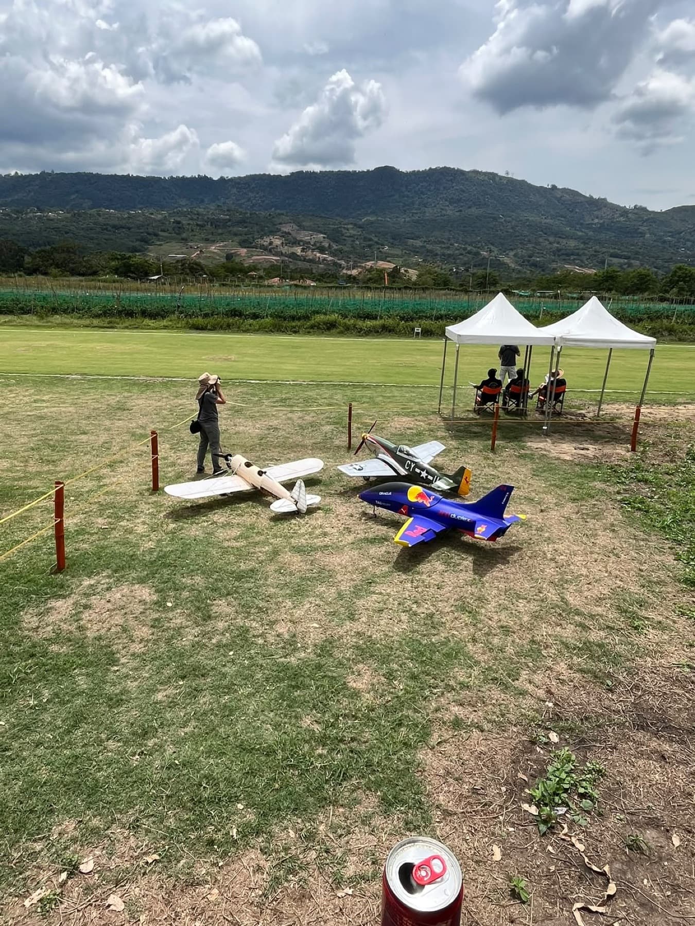 Aviones en el césped dentro de una zona delimitada con estacas y cuerda; al fondo, la pista y una carpa con jueces.