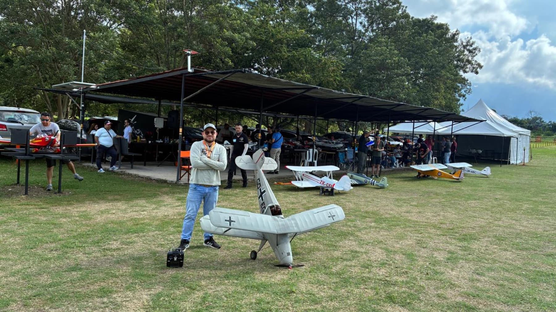 Un piloto de pie junto a un aeromodelo de ala alta frente a los pits del club.