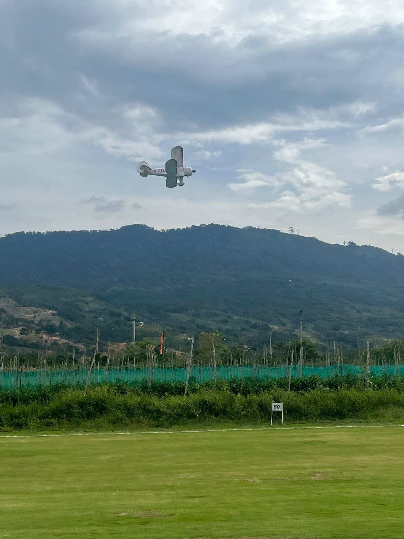 Biplano a escala en vuelo sobre el campo, con la montaña y las nubes de fondo.