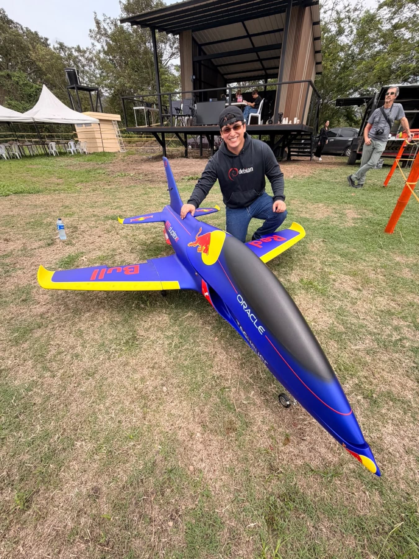 Piloto sonriente agachado junto a un jet a escala azul con puntas amarillas, sobre el pasto del campo.